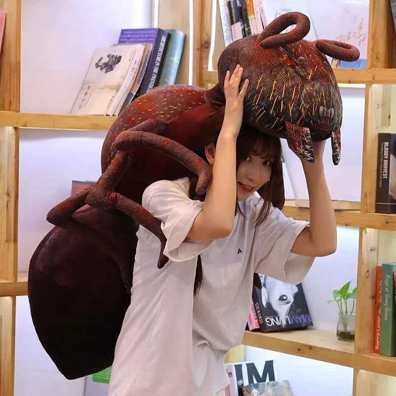 A giant 100cm realistic red ant plush toy with long legs and antennae, displayed as a decorative pillow on a modern white bed.