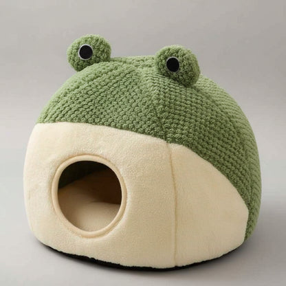 Cat lying inside a green and white frog-shaped pet bed on a wooden floor.